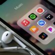 iPhone com EarPods e vários apps de música: Apple Music, Spotify, YouTube Music, Shazam, TIDAL, Deezer, Pandora e mais