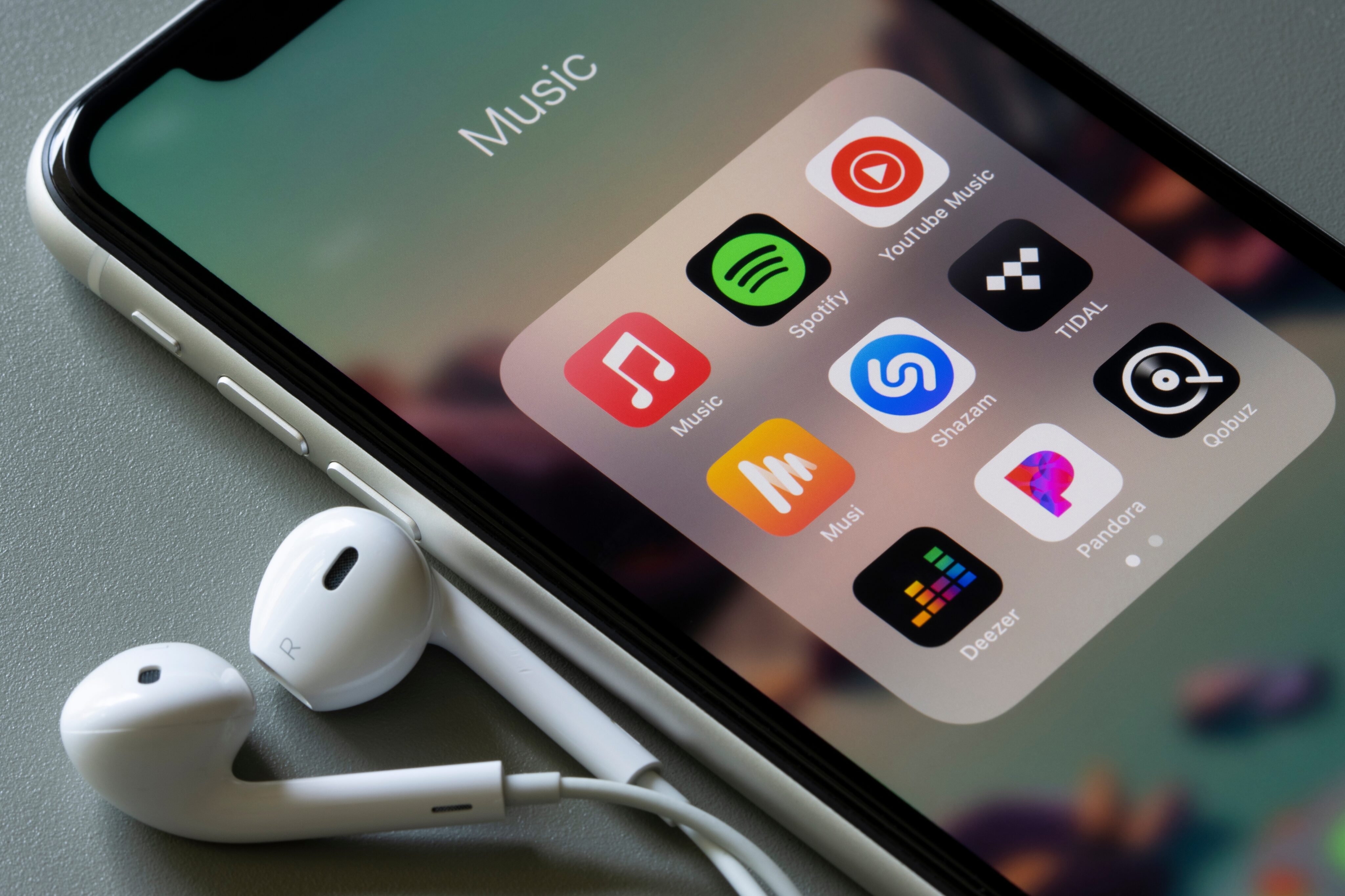 iPhone com EarPods e vários apps de música: Apple Music, Spotify, YouTube Music, Shazam, TIDAL, Deezer, Pandora e mais