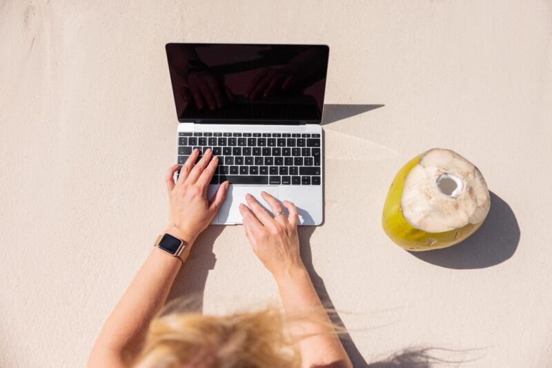 Mulher trabalhando em MacBook numa praia com côco ao lado