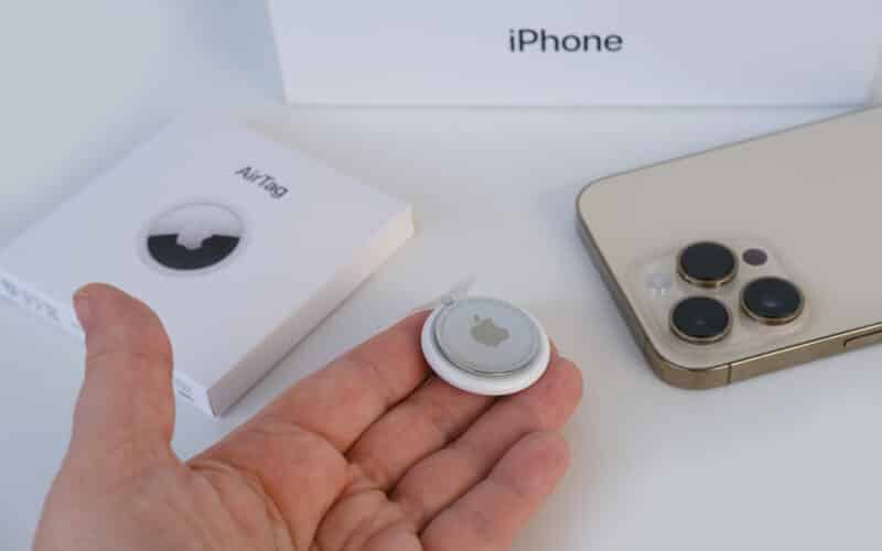 Homem com um AirTag na mão; ao lado, um iPhone, uma caixa de AirTag e uma caixa de iPhone