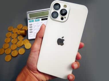 iPhone Pro Max em mão com moedas e calculadora ao fundo