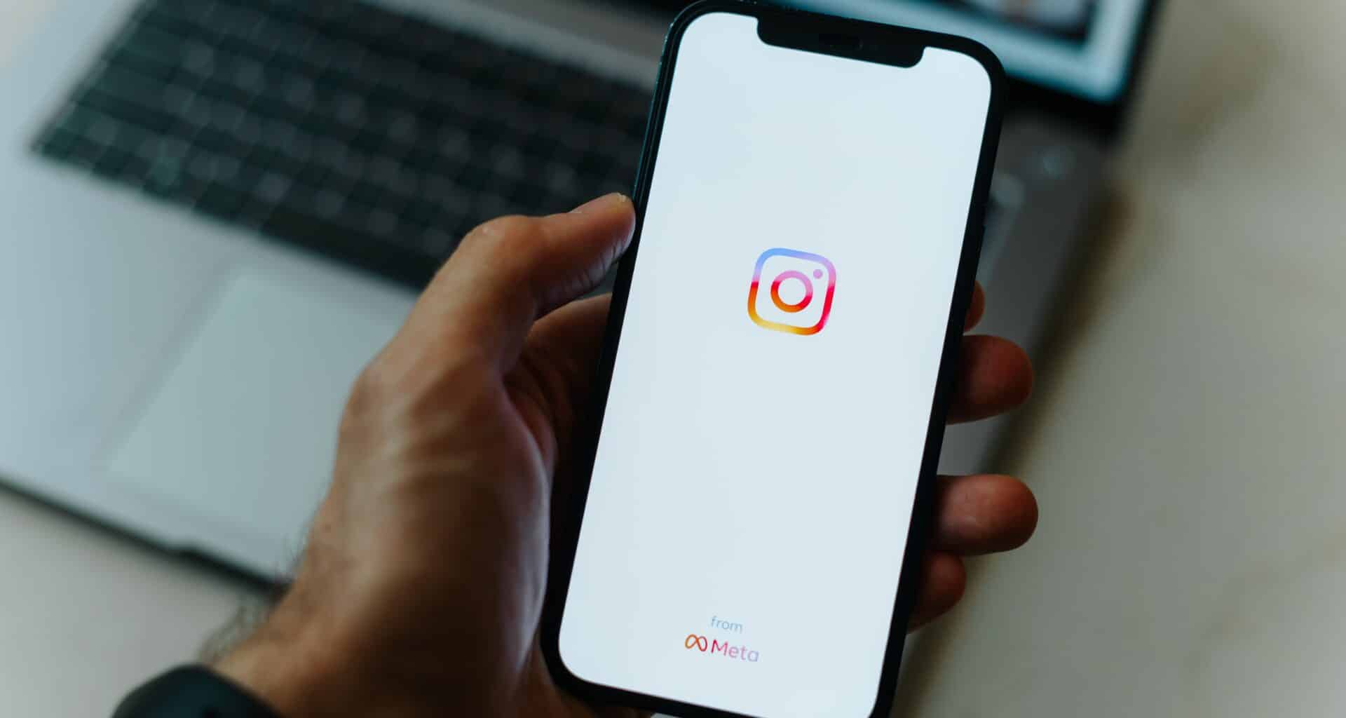 Mão segurando o iPhone com o Instagram com um Mac ao fundo
