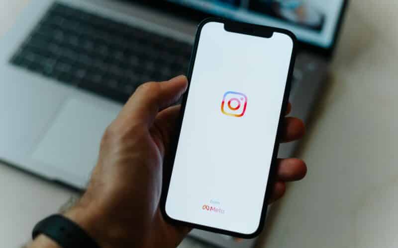 Mão segurando o iPhone com o Instagram com um Mac ao fundo
