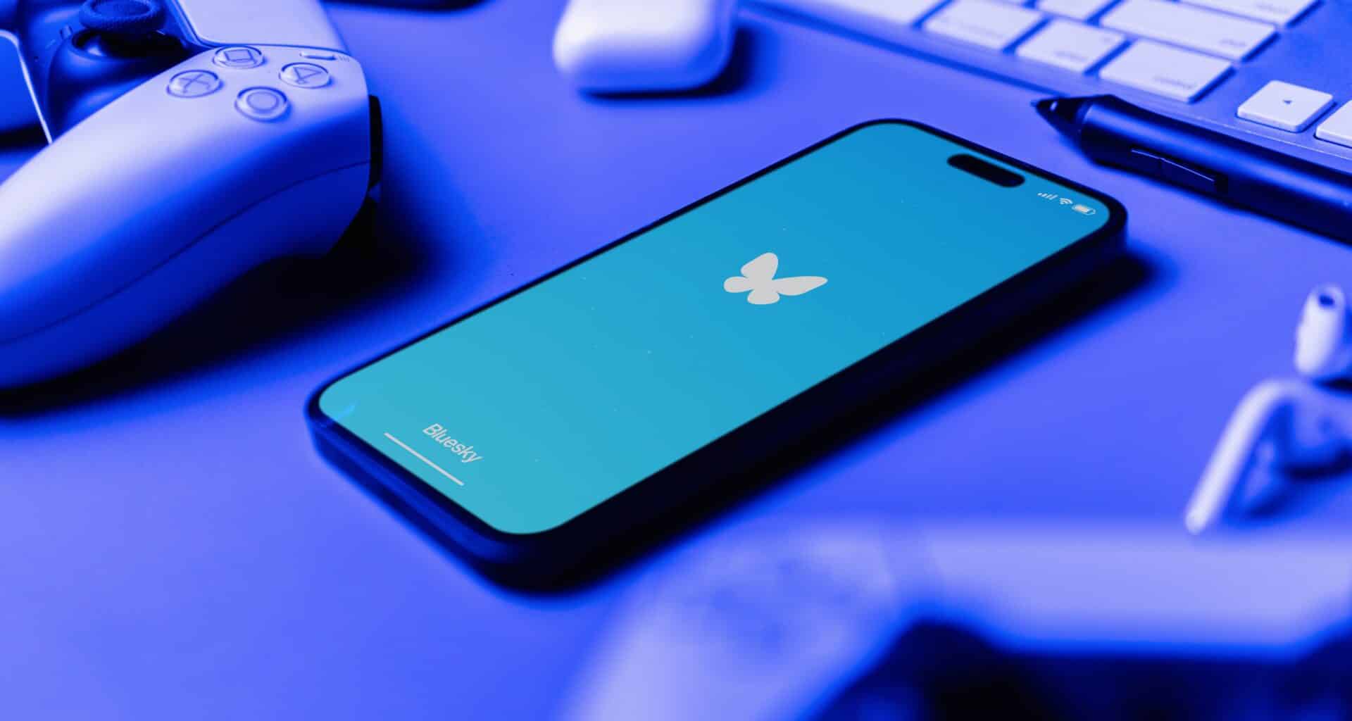 App Bluesky no iPhone ao lado de um controle de PS5, AirPods e teclado