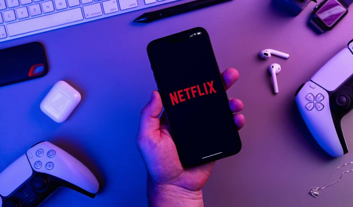 App da Netflix aberto do iPhone com diversos acessórios (AirPods, teclado, controles do PS e mais) ao lado