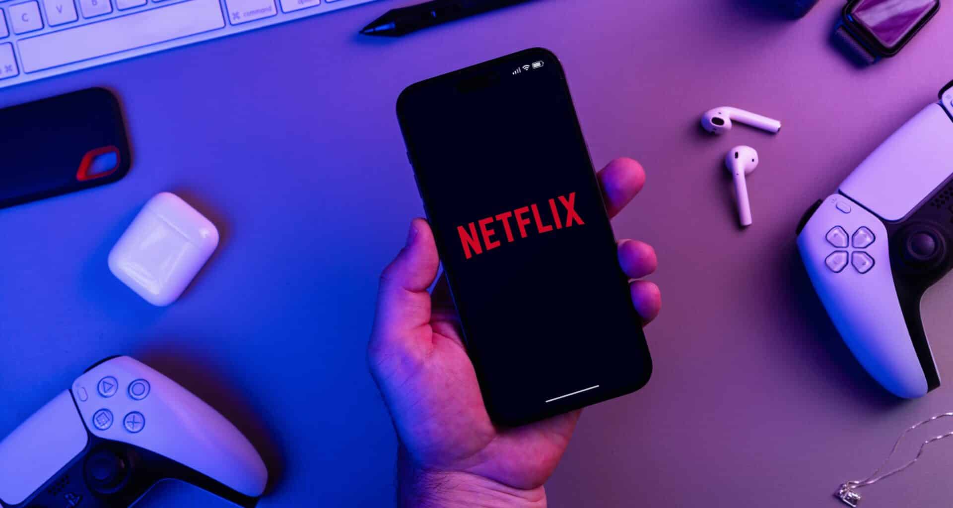 App da Netflix aberto do iPhone com diversos acessórios (AirPods, teclado, controles do PS e mais) ao lado