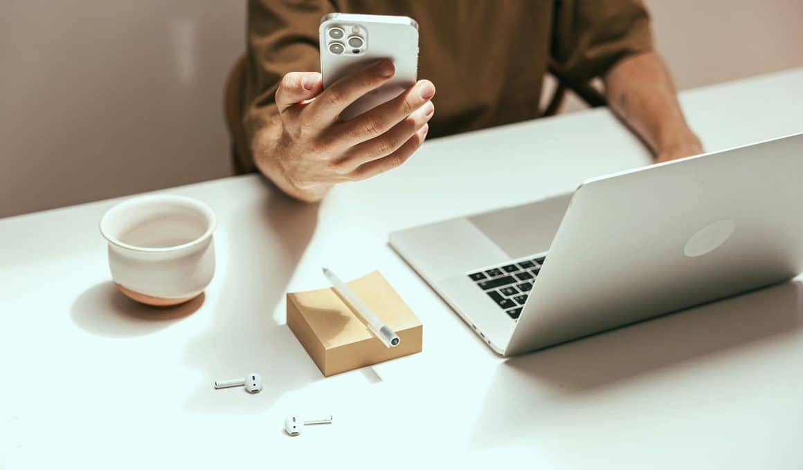 Homem usando um iPhone com um Mac e AirPods em cima da mesa