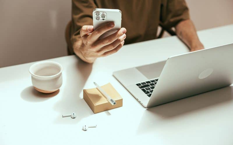 Homem usando um iPhone com um Mac e AirPods em cima da mesa