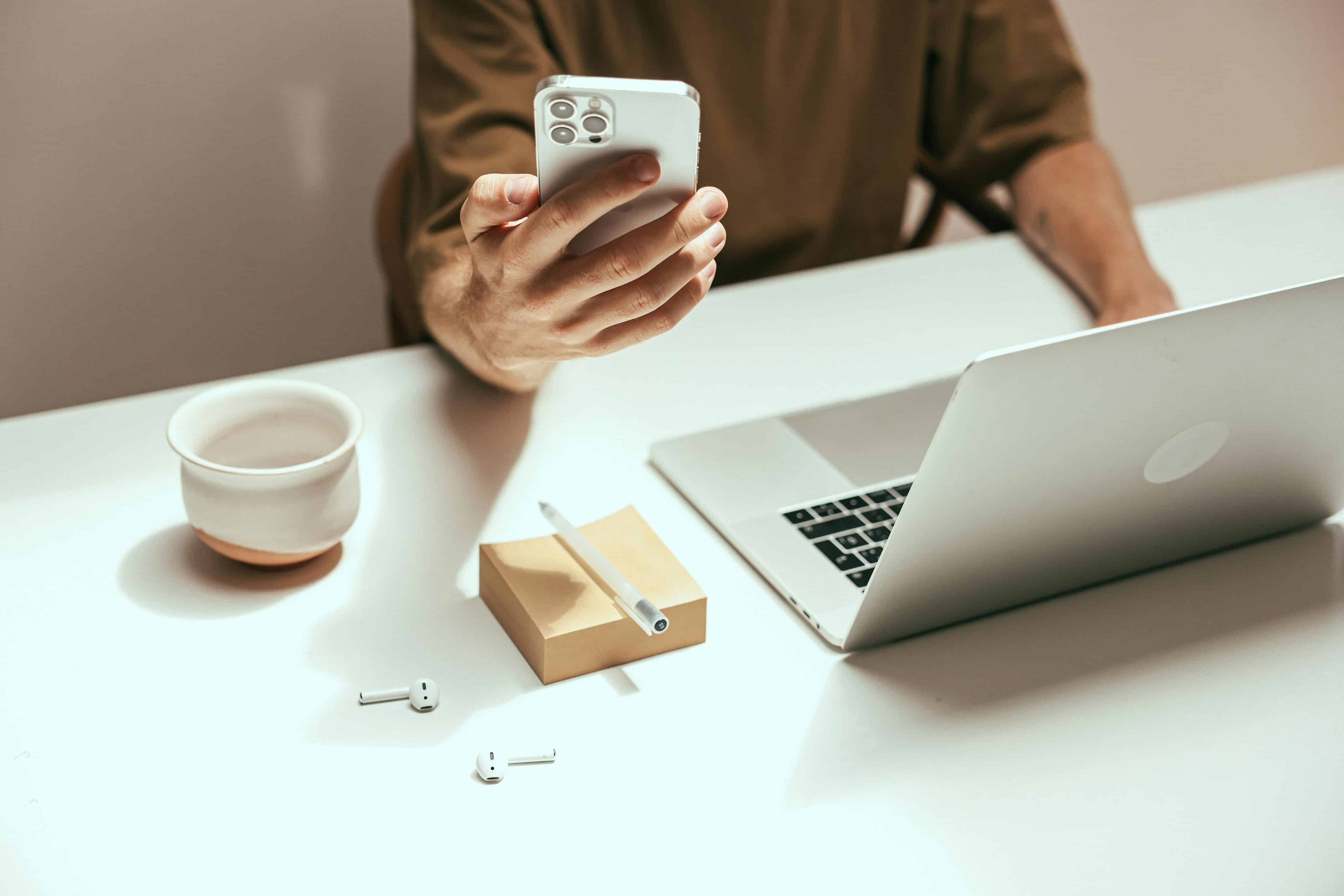 Homem usando um iPhone com um Mac e AirPods em cima da mesa