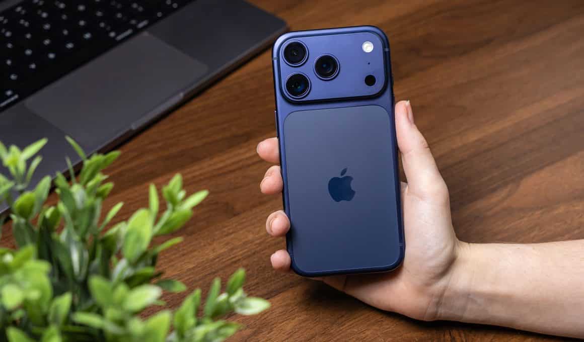 Mão segurando um iPhone 17 Pro azul com um MacBook Pro ao fundo